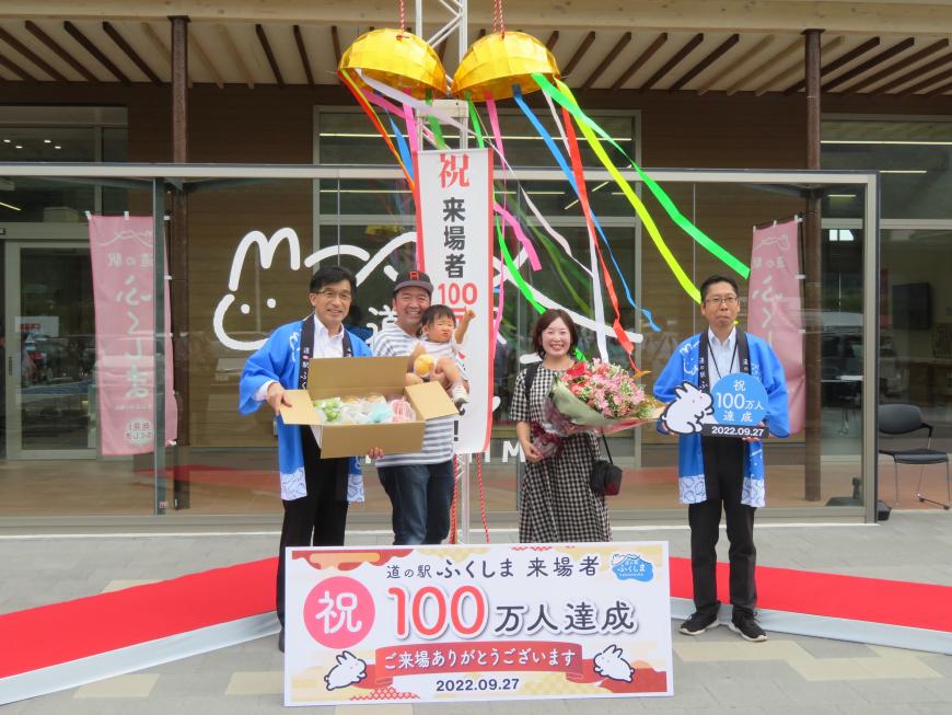 お祝いのくす玉が割られた前で記念品が入った段ボールを持った市長と100万達成のボードを持った男性の間に子どもを抱っこした父親と花束を抱えた母親が立っている写真
