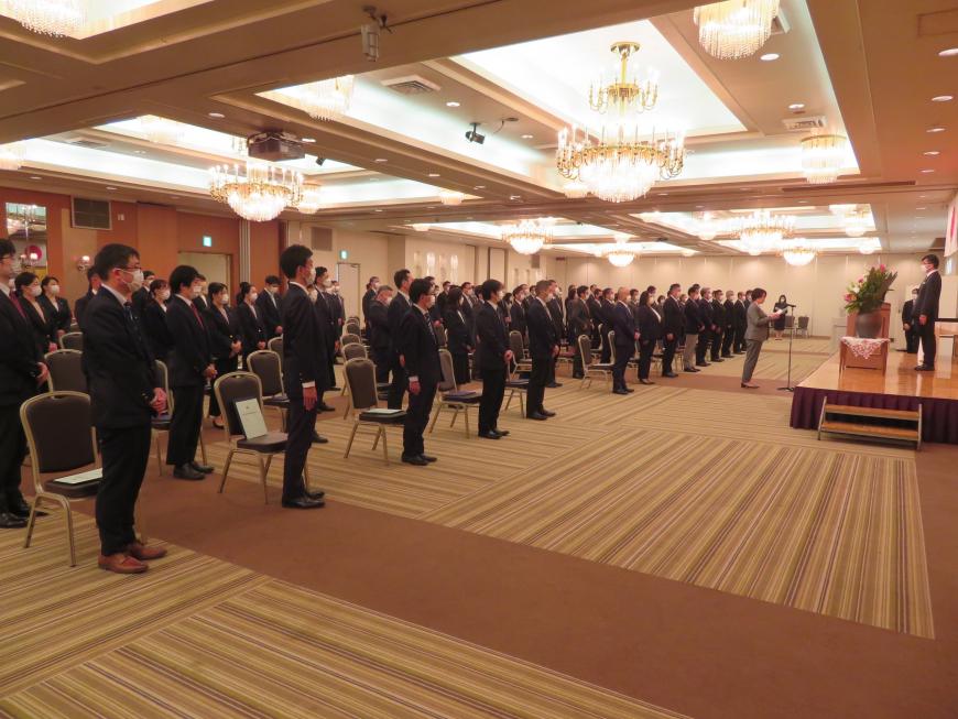 大勢の出席者が起立して姿勢を正し、代表の人がステージの上に立った市長の前で挨拶をしている写真