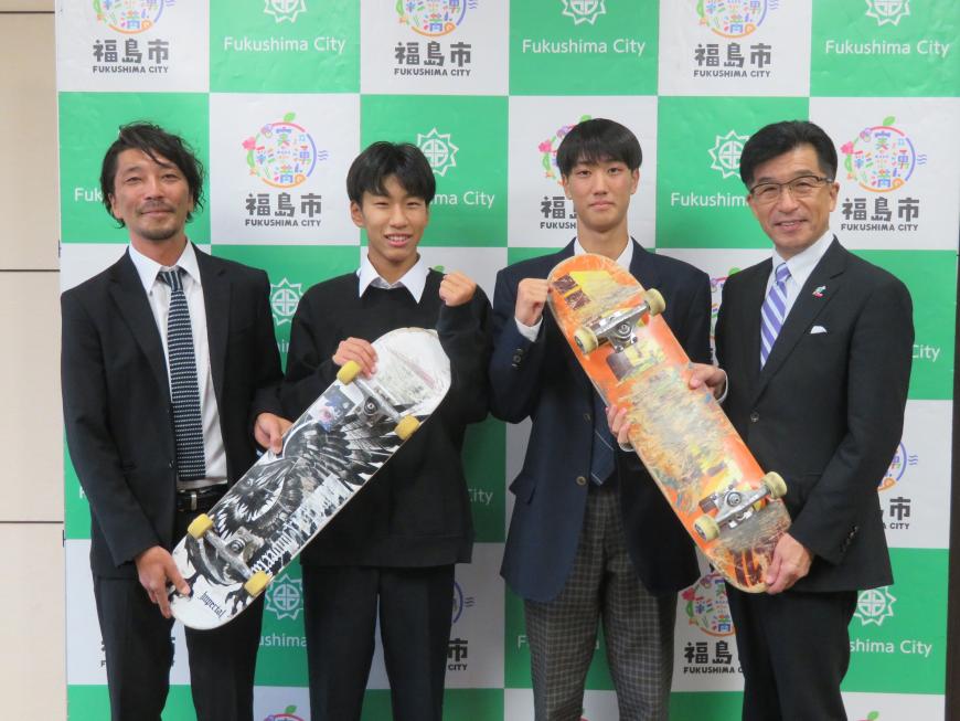 バックパネルの前に市長を含む男性4名が立ち、2名で1つのスケートボードを持っている写真