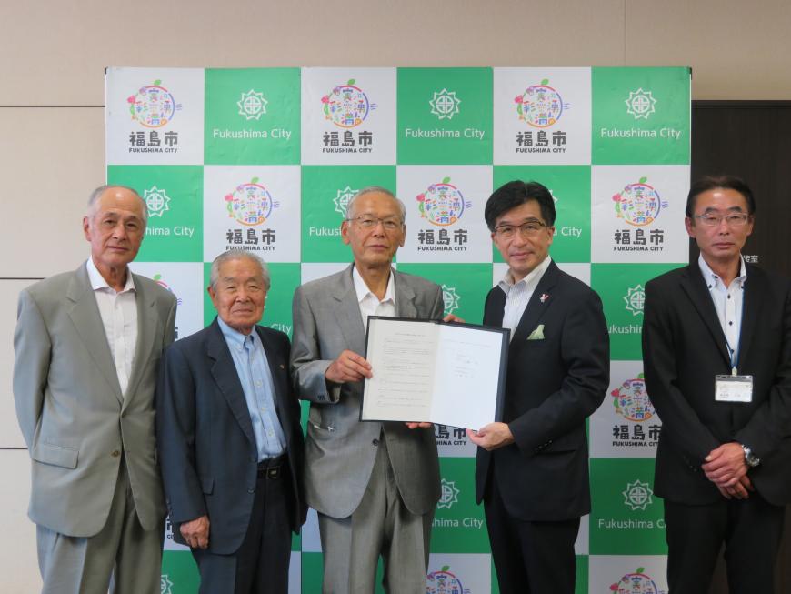 バックパネルの前で、市長と男性で協定書を持ち、他の男性が2名と1名に分かれて横に立っている写真