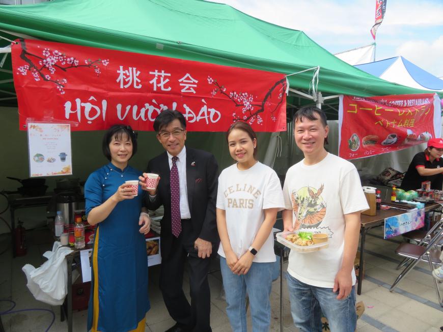 「桃花会」と書かれた幕が吊るされたテント前で市長と女性が紙コップを持ち、男性が料理が盛られた皿を持っている写真