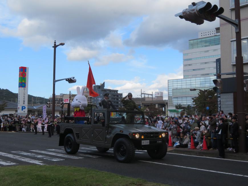 沿道で多くの人が見ている中、敬礼をした自衛官を乗せた車両がパレードしている写真