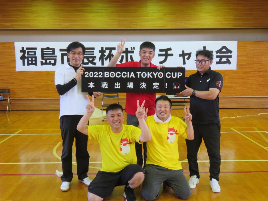 体育館で黄色のTシャツを着た男性2名が中腰でピースサインをしている後ろで市長と男性2名で本戦出場決定のボードを持って立っている写真
