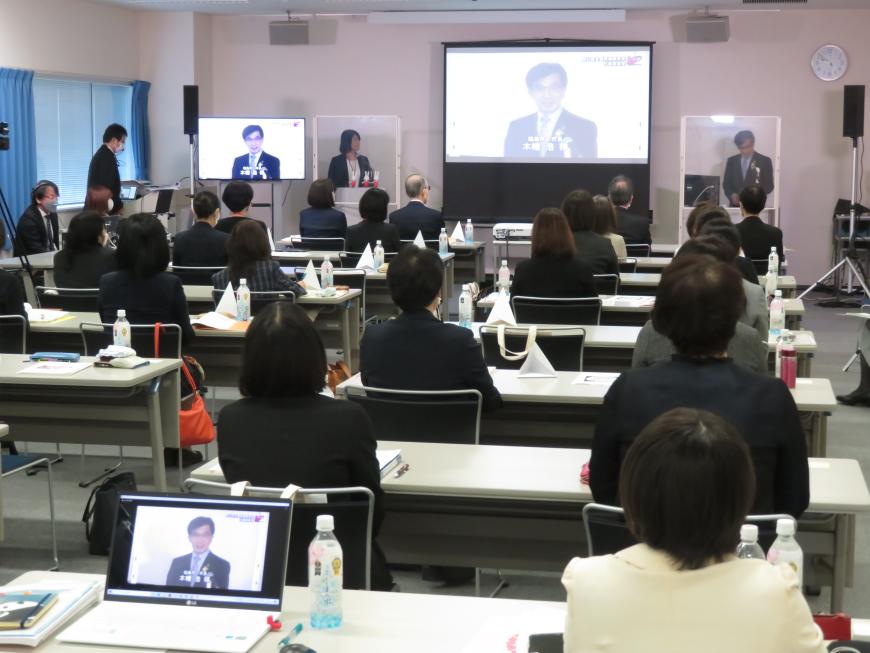 長机に2人ずつ座っている参加者の前でアクリル板で仕切られた演台に立って市長が話をしており、大きなスクリーンやモニターに市長が映し出されている写真