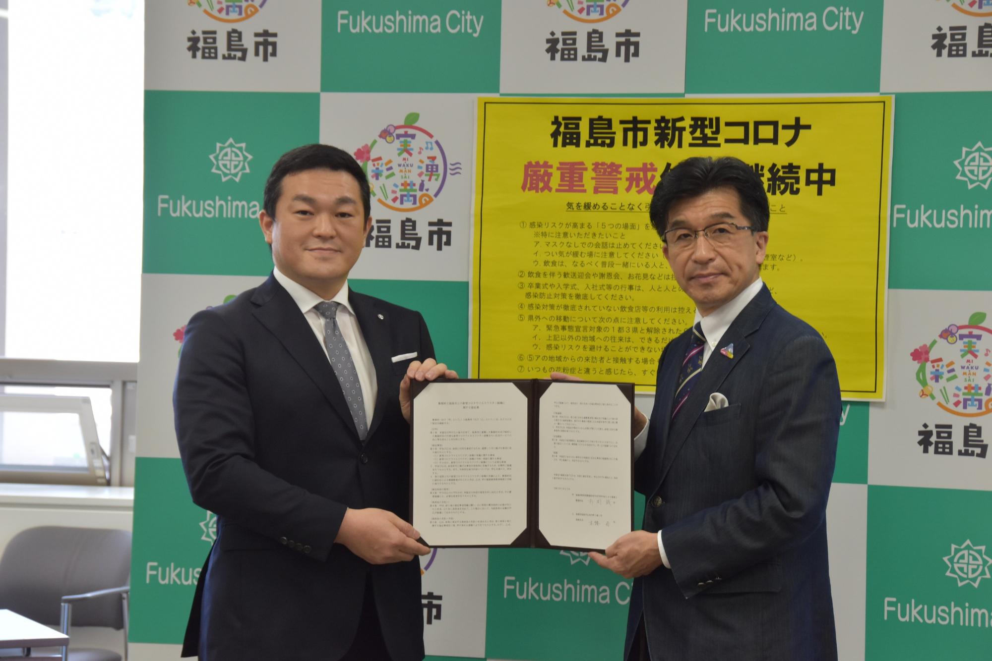新型コロナ厳重注意の紙が貼られたバックパネルの前で、市長と男性で協定書を持って立っている写真