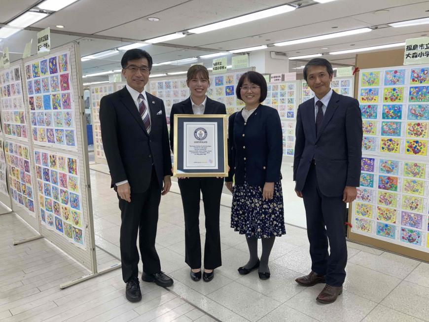 数多くの塗り絵が飾られた会場で、市長と認定書を持った女性、関係者の男性と女性が並んでいる写真