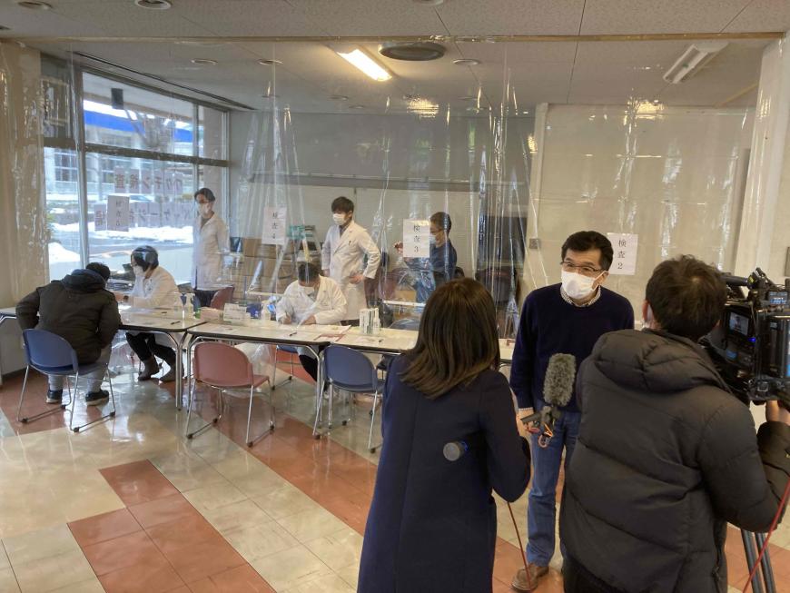 透明のビニールシートで区切られた奥に白衣を着た人がいる会場で、市長がテレビカメラの前で女性からの取材を受けている写真