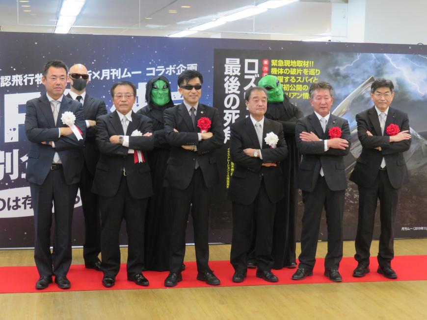 大きな展示ポスターの前で、前列にサングラスをした市長と男性5人、後列に宇宙人に仮装した2人が立ち、腕組みをして撮影した写真