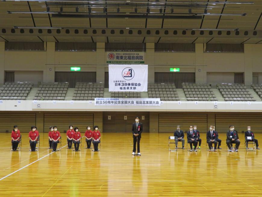 日本3B体操協会の横断幕が吊るされた体育館で、立って話をしている市長と、その左右に並んだ椅子に座ったお揃いの赤のポロシャツ姿の女性たちと関係者の男性の写真