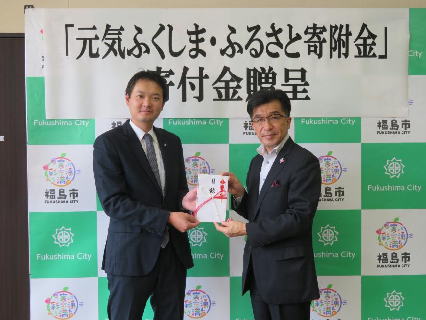 寄附金贈呈の横断幕が貼られたバックパネルの前で、市長と男性で目録封筒を持っている写真
