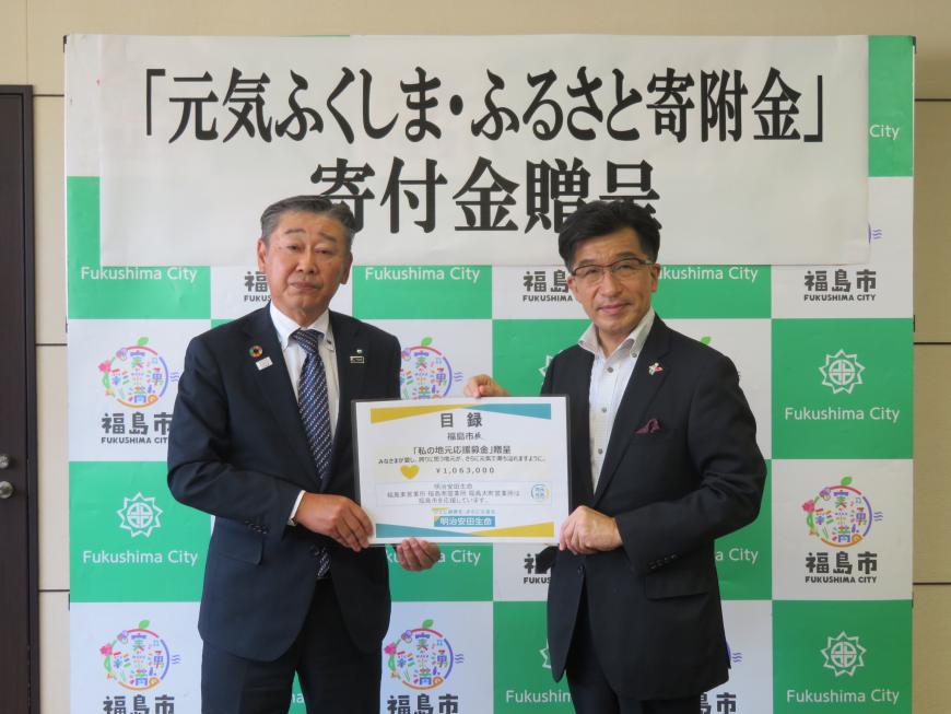 寄附金贈呈の横断幕が貼られたバックパネルの前で、市長と男性で目録のボードを持っている写真