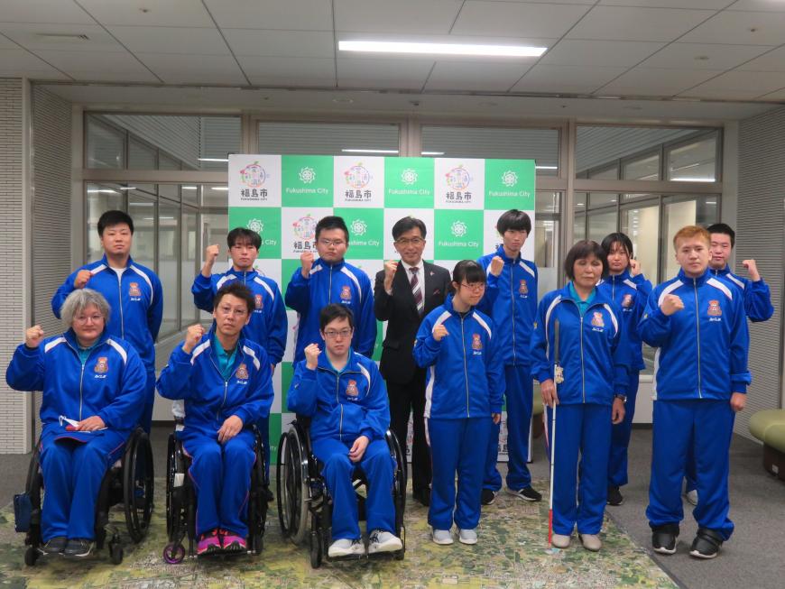 車いすに乗った3名を含む12名の選手がお揃いの青のジャージ姿で市長と2列に並んでガッツポーズをしている写真