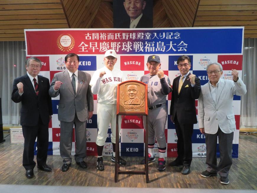 古関裕而の顔が描かれた盾の後方で早稲田のユニフォーム姿の男性の右隣に男性2名、慶應のユニフォーム姿の男性の左隣に市長と男性が立ちそれぞれ片手でガッツポーズをしている写真