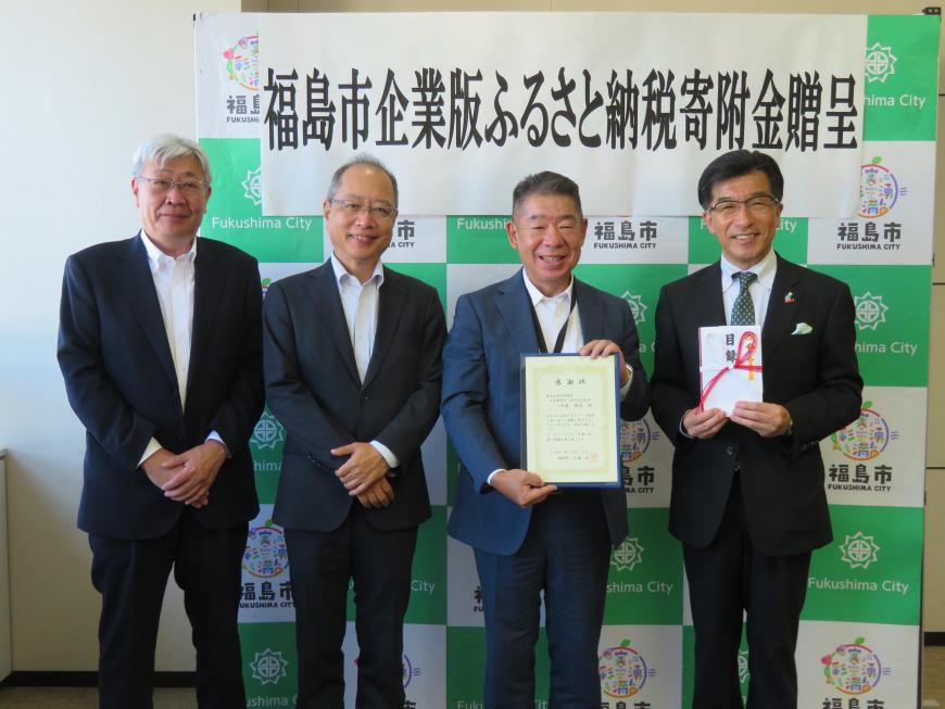 「福島市企業版ふるさと納税寄附金贈呈」の幕が貼ってあるバックパネルの前に市長を含む男性4名が立ち、市長が目録、男性1名が額入りの賞状を持っている写真