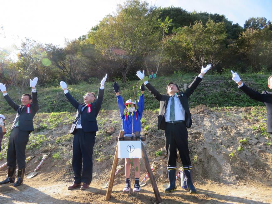 1番の札がつけられた苗木の後ろで児童と市長と関係者の方が横一列に並んで空に向かって手を広げている写真