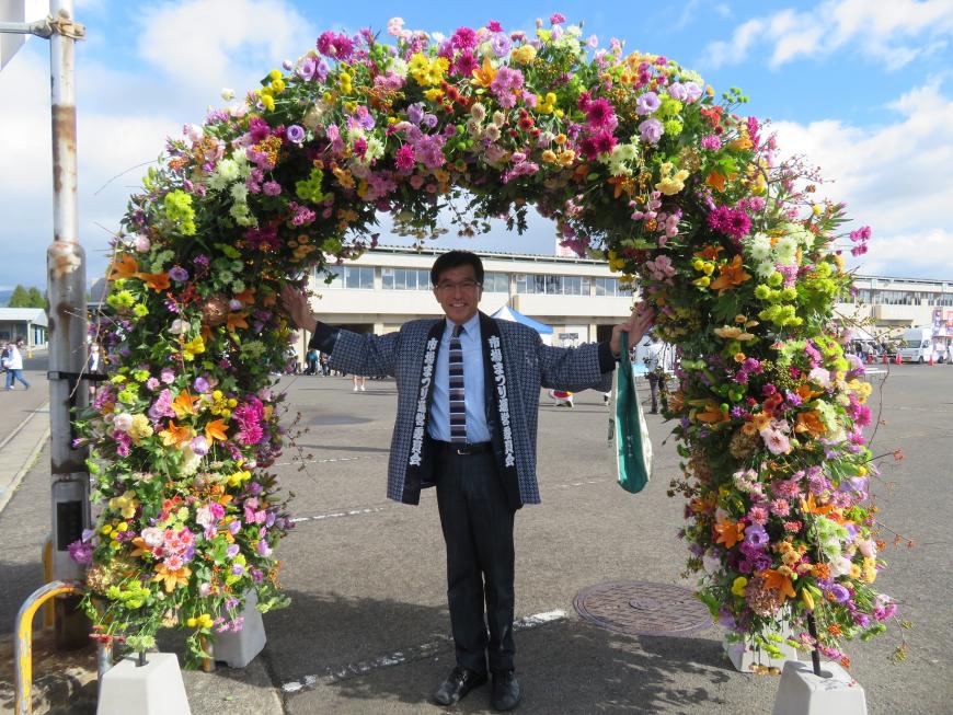花で装飾されたゲートで法被を着た市長が両手を広げている写真