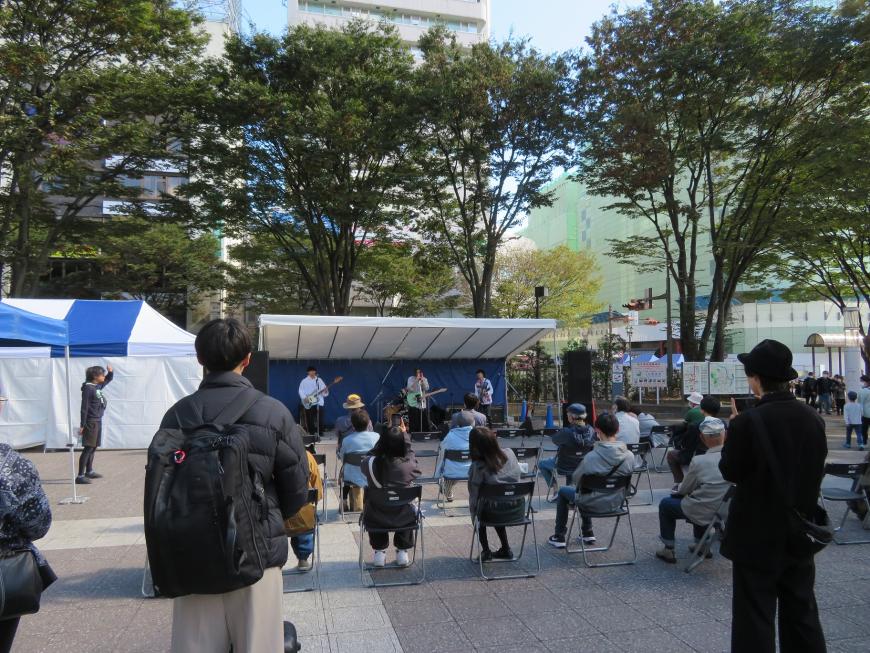 屋外の屋根付きステージで3名のバンドの演奏を観客が聞いているところを後方から撮影した写真