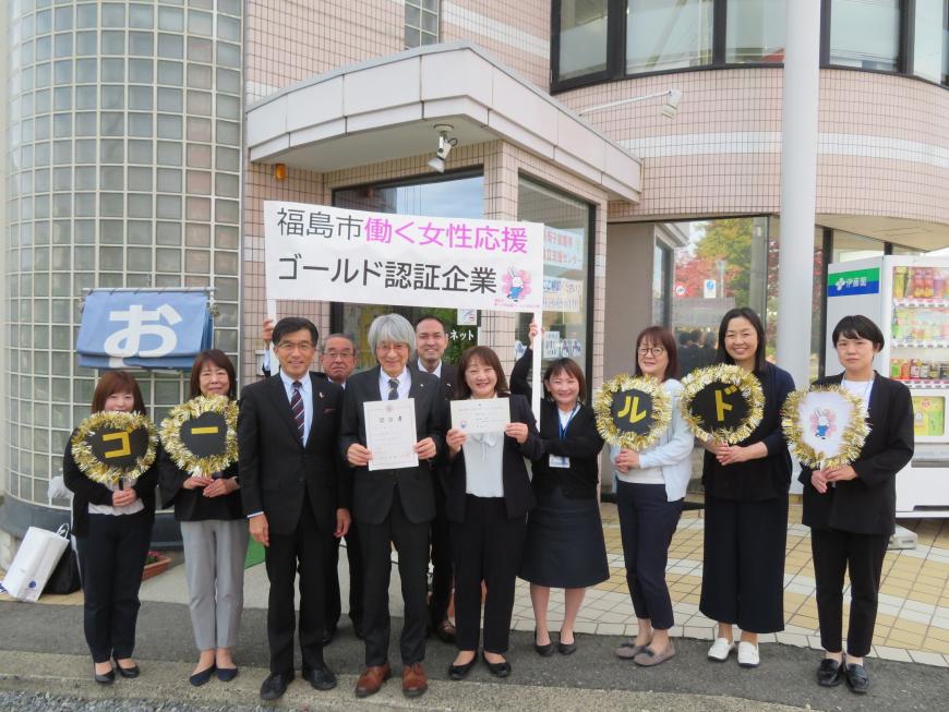 建物入口で「福島市働く女性応援ゴールド認定企業」のプラカードの前で認定書や一文字ずつゴールドと装飾したうちわを持った企業の方々と市長が集まって撮影した写真