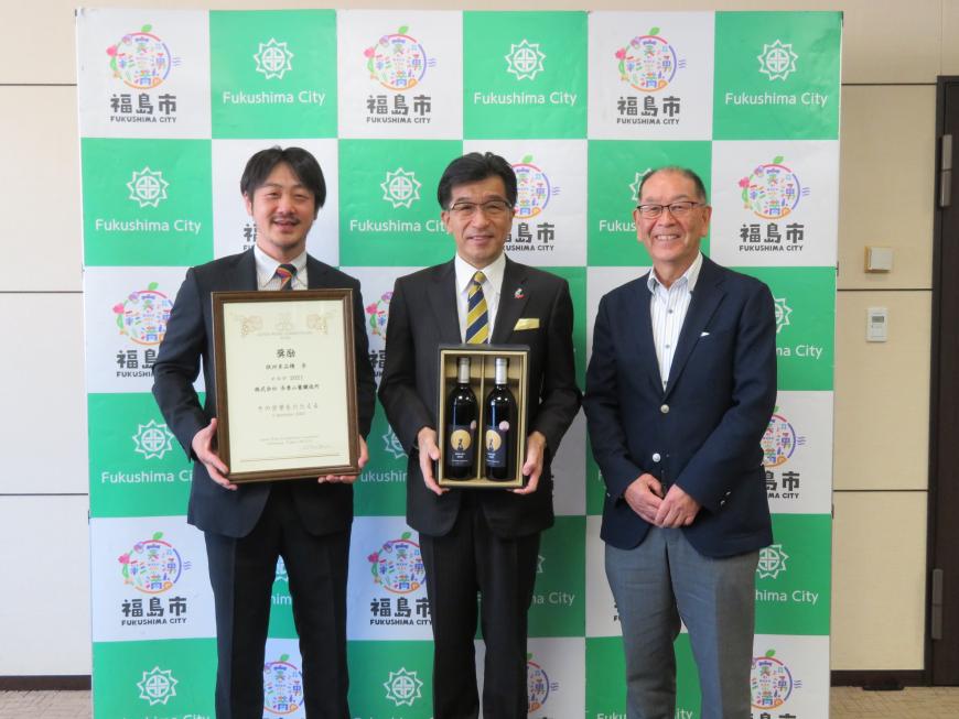 バックパネルの前に市長の両脇に男性2名が立ち、市長がワインボトルが2本入った箱、左隣の男性が額入りの賞状を持っている写真