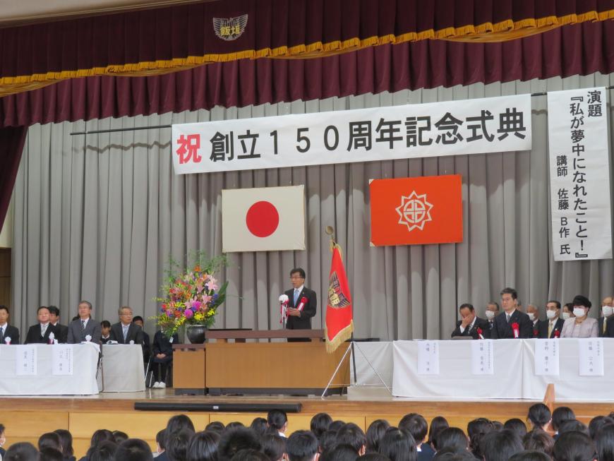 「創立150周年記念式典」の横断幕があるステージに来賓の方々が座っている中で演台で市長が話をしている写真
