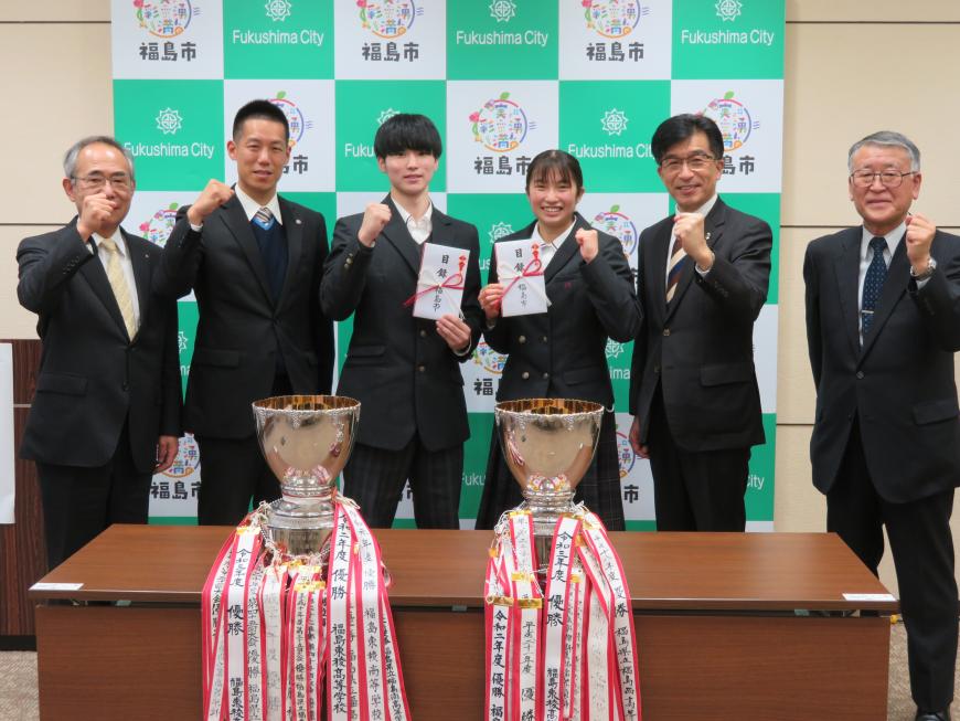 優勝カップ2個を前にして、市長、目録封筒を持った男子生徒、女子生徒と関係者3人がガッツポーズをしている写真