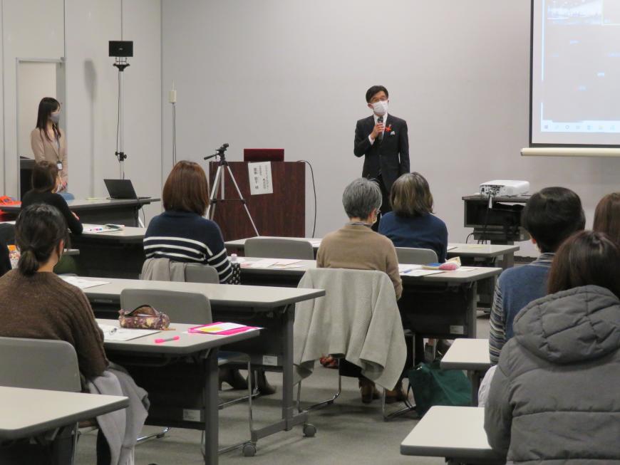 長机の席に出席者の女性たちが2人ずつ座っている前で、市長が立って話をしている写真