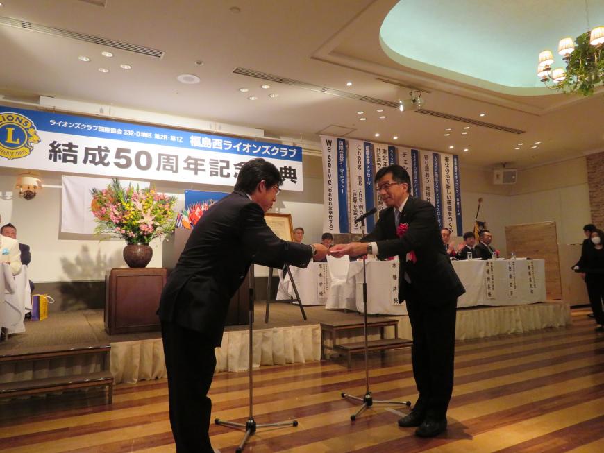「結成50周年記念式典」の横断幕があるステージに来賓の方々座っている前で市長が男性に目録を渡している写真