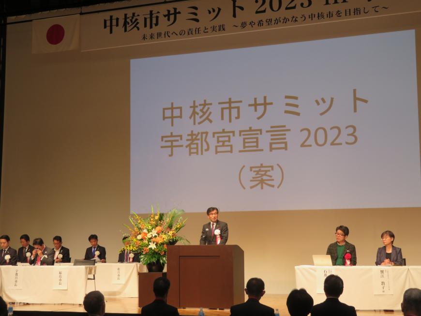 スクリーンに「中核市サミット宇都宮宣言2023(案)」と映し出されたステージで来賓の方が座っている中、演台で市長が話をしている写真