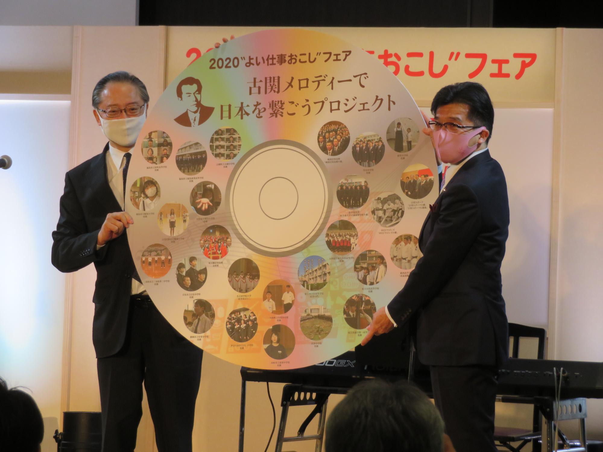 「2020”よい仕事おこし”フェア 古関メロディーで日本を繋ごうプロジェクト」と記された大きなDVDのレプリカを、市長と関係者が両手で抱えている写真