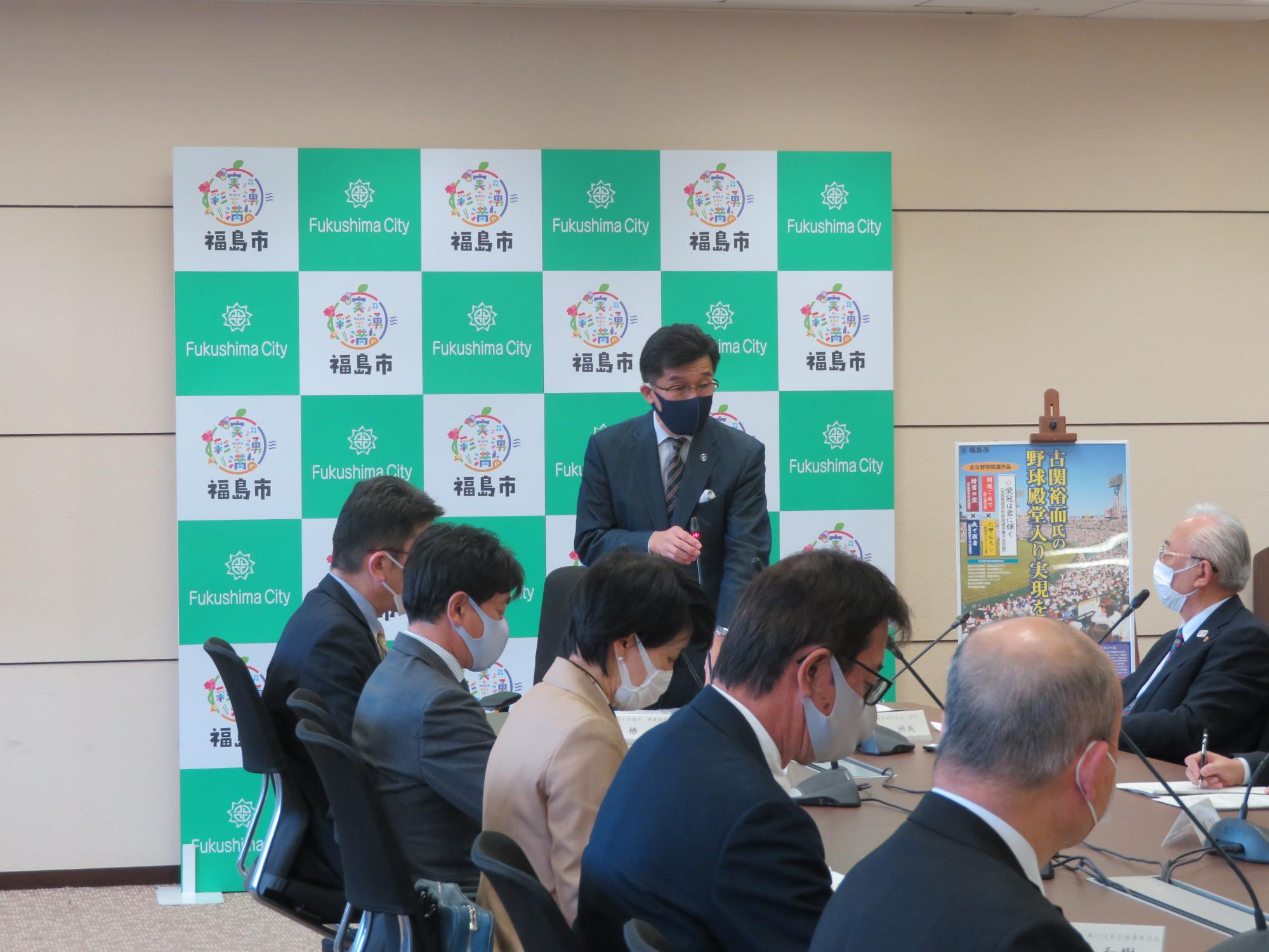 福島市のバックパネル前で、市長が席に座っている関係者に向けて話をしている写真