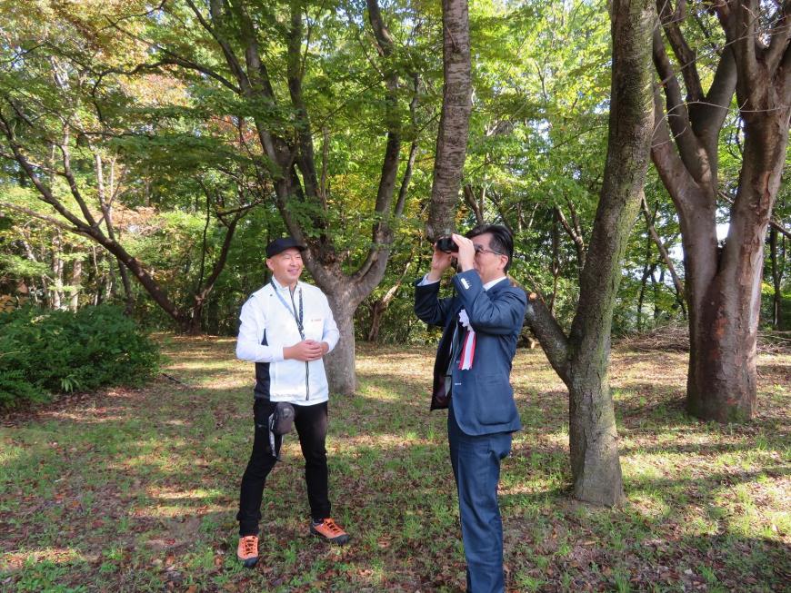 陽が差し込んでいる森の中で、職員が立っている横で市長が双眼鏡で遠くをみている写真