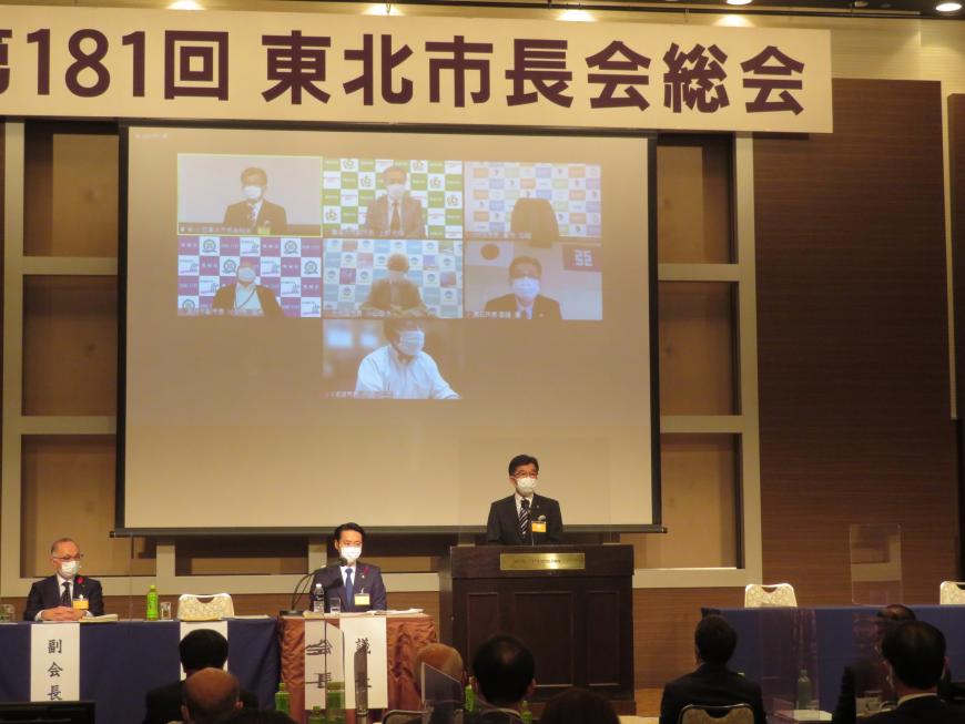 ステージのモニターにネットでつながった他の参加者が映し出される中、市長がステージの演台で話をしている写真