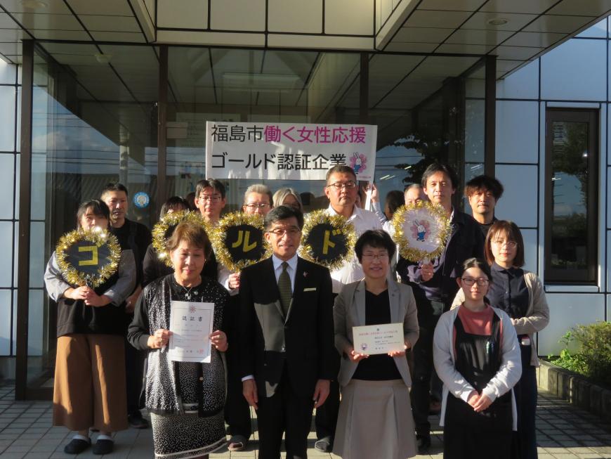 「福島市働く女性応援ゴールド認証企業」の幟がある建物入口で認定書を持った女性の間に市長が立ち、その後列にゴールドの文字を一文字ずつ装飾したうちわを持った関係者が並んでいる写真