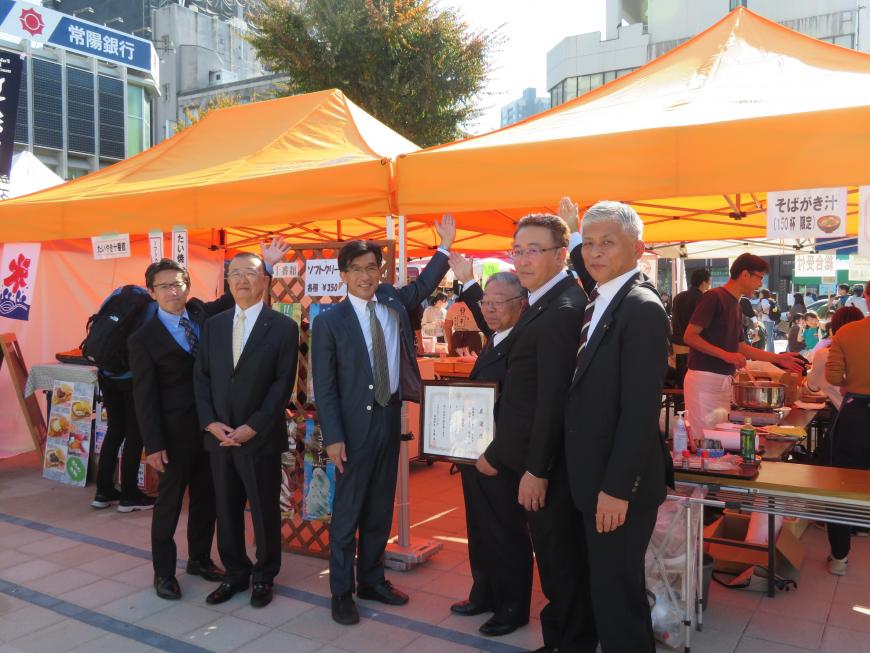 屋外のオレンジ色のテントに前に市長と男性5名が立ち、1人が額入りの賞状を持ち、テントにそれぞれ手を向けている写真