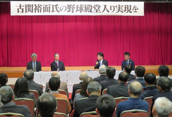 「古関裕而氏の野球殿堂入り実現を」の吊りパネルの下、ステージ前で市長と関係者が席に座っており、参加者が椅子に座り話に耳を傾けている写真