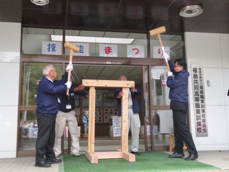 建物の入り口で男性2名が支えている角材に、市長と男性がそれぞれ木槌を持って、他の角材を打ち込んでいる写真