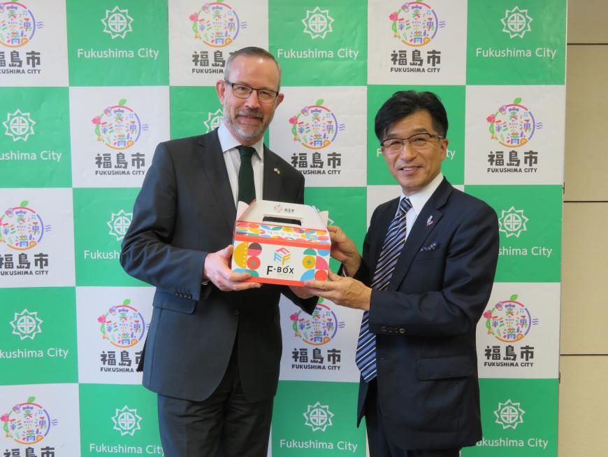 バックパネルの前で市長が駐日大使にFBOXを手渡している写真