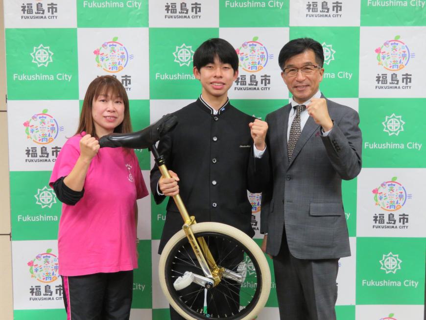 バックパネルの前に市長と女性の間に制服姿の男子生徒が一輪車を持って立ち、それぞれガッツポーズをしている写真