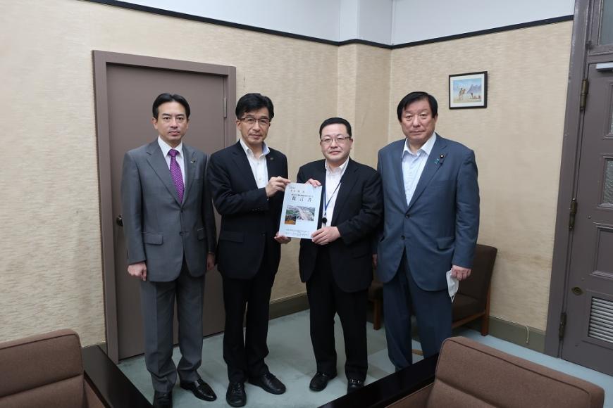 応接椅子が置かれた部屋の中で市長と男性が要望書に手を携えて立ち、その両脇にスーツを着た男性がそれぞれ1人ずつ立って写った記念写真