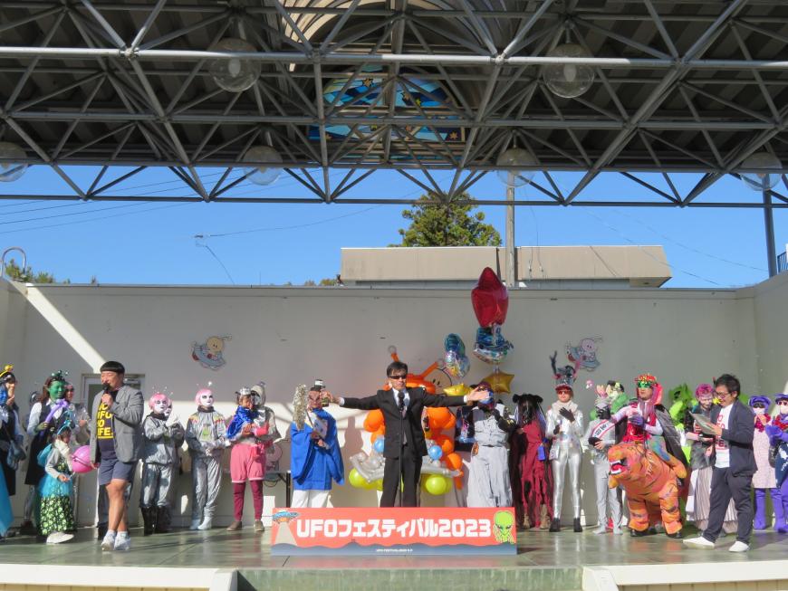 ステージ上に仮装した多く人がいる中、サングラスかけた市長が中央で両手を広げている写真