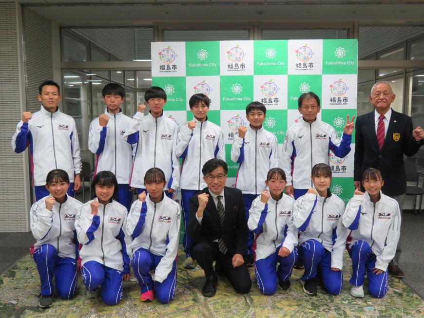 バックパネルの前で、前列は市長と女子選手がひざまづいて、後列は男子選手と関係者が立ってそれぞれがガッツポーズをしている写真
