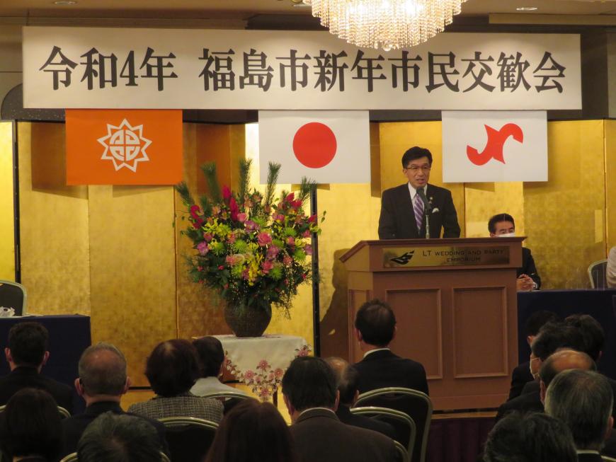 福島市新年市民交歓会のボードがある会場で、座っている多くの参加者の前で、市長が金屛風の前にある演台に立って話をしている写真