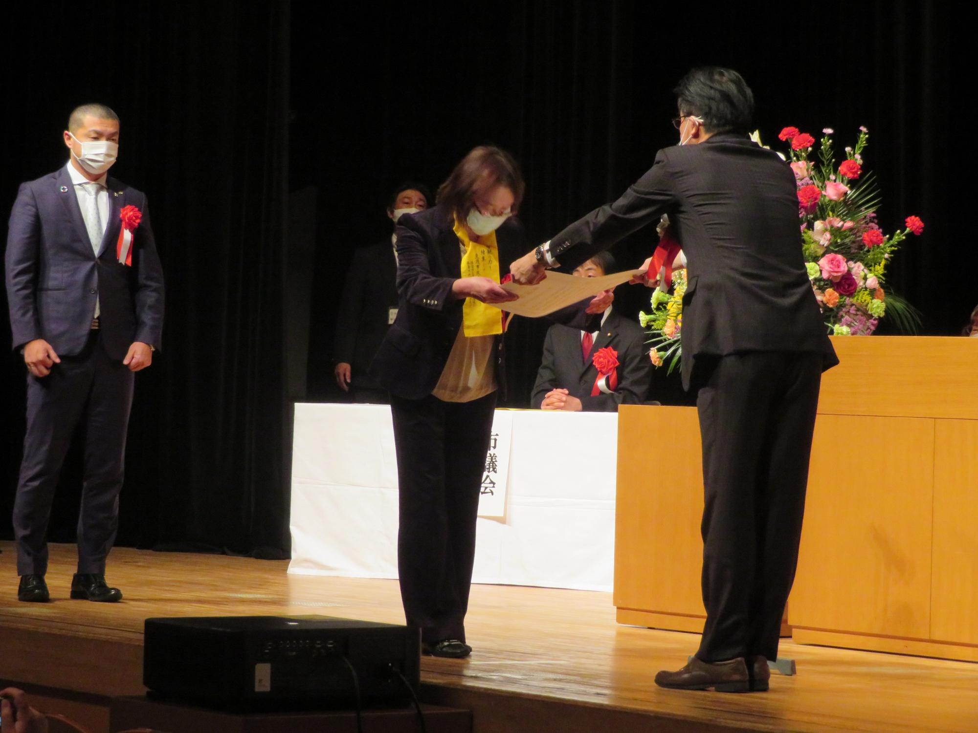 市長がステージの演台前で、女性の表彰者へ感謝状を手渡している写真