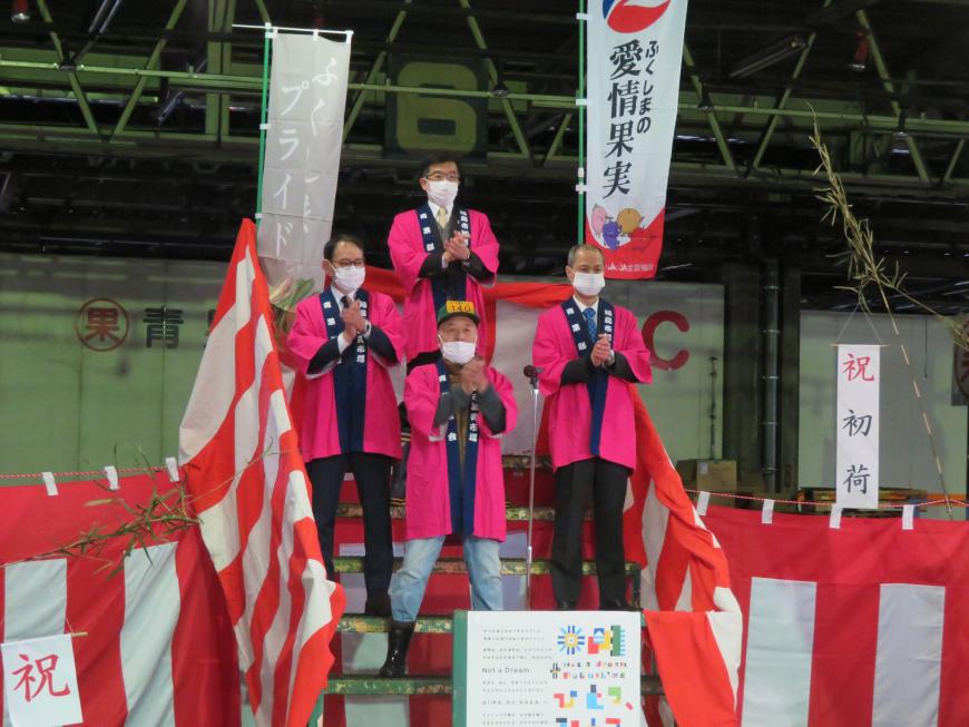初荷の幟が立っている会場で階段にピンクの法被を着た市長を含む男性4人がひし形に並んで立ち、手を合わせている写真