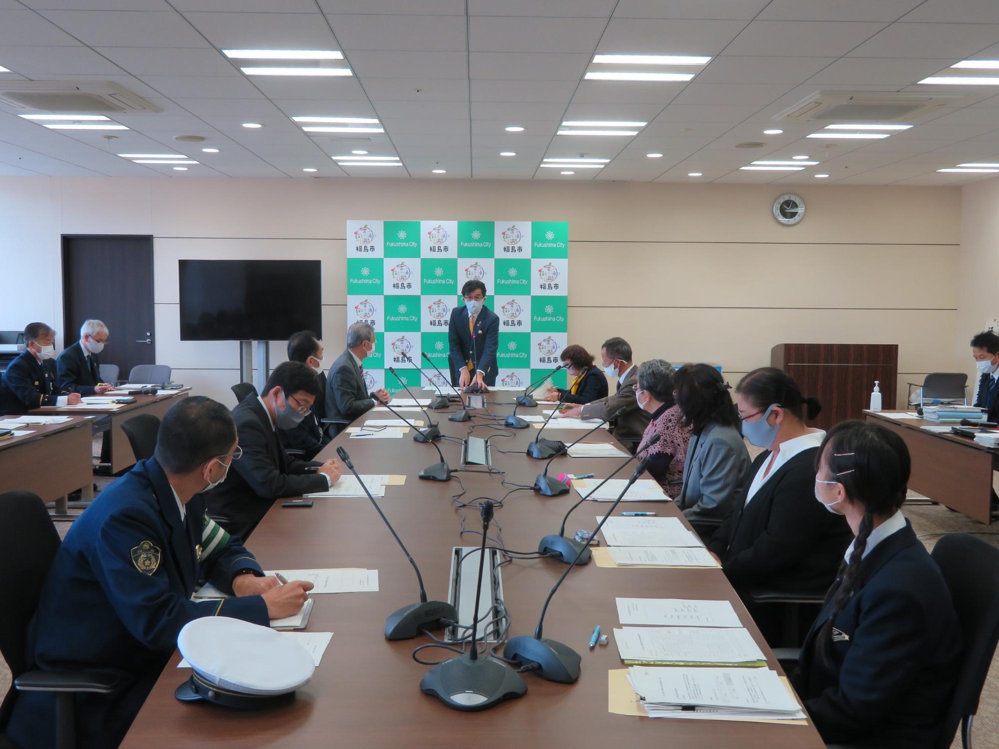 庁議室の壁に設置された福島市のバックパネル前に立っている市長に、長机に座った出席者たちが目を向けている交通安全対策の会議の様子の写真