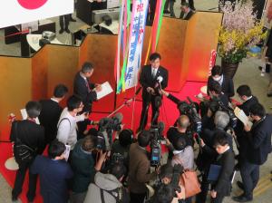 金屛風の前でくす玉の垂れ幕の前に立った市長がたくさんの取材陣から取材を受けている様子を上から写した写真