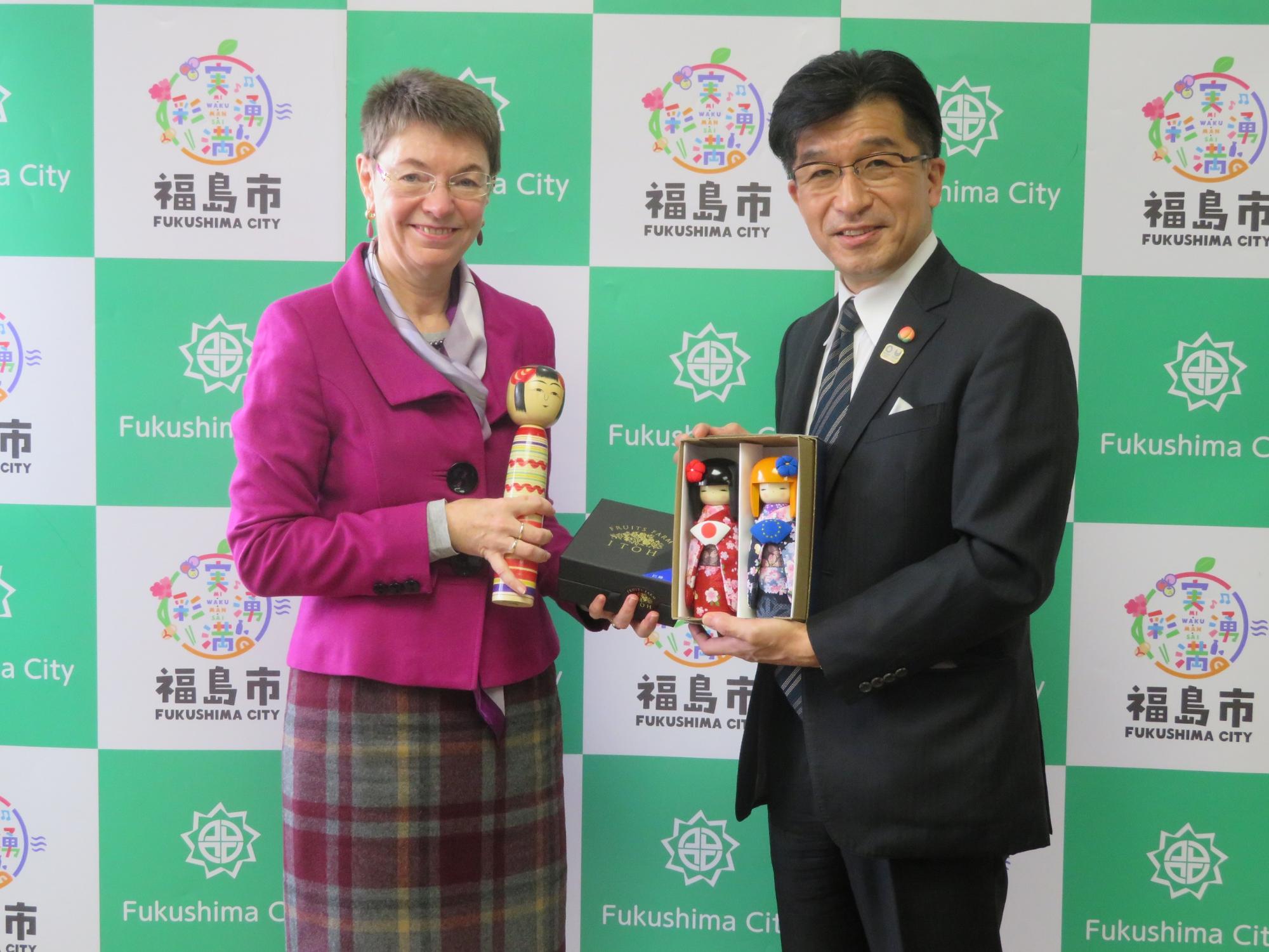 福島市のバックパネル前で、市長が箱に入った人形を手に持ち、駐日欧州連合特命全権大使の関係者の女性はこけしを持っている写真