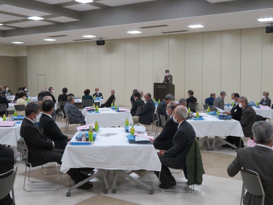 1つのテーブルに4人ずつに分かれて座っている参加者の前で、市長が演台に立って話をしている写真