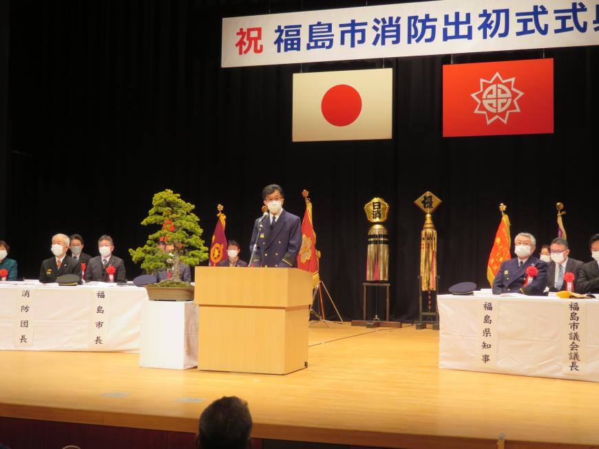 「福島市消防出初式」のボードがある会場で、市長が来賓の方が座っているステージの演台に立って話をしている写真