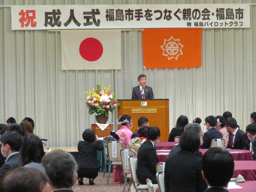 成人式の横断幕がある会場で、円卓テーブルに分かれて座っている参加者の前で、市長が演台に立って話をしている写真